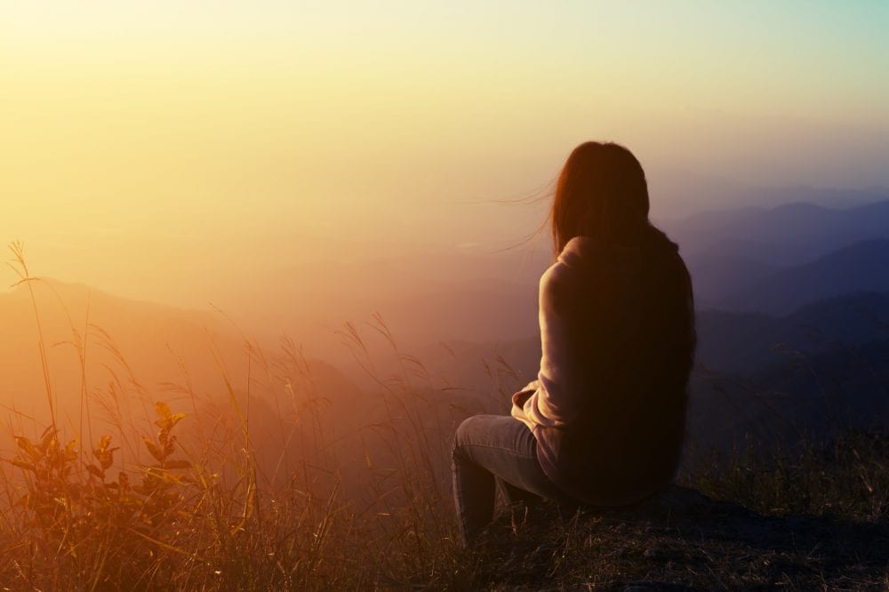 Woman sitting on a rock, looking out over a beautiful view on a mountain representing The First Step in Recovering From a Breakup: Validation.