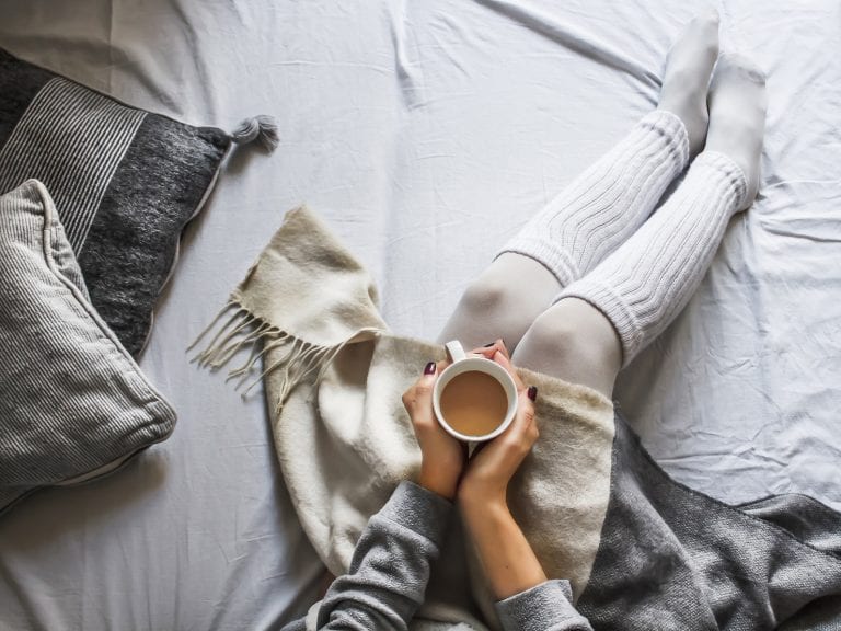 girl sitting in bed with heartbreak over the holidays holding a cup of tea representing How to Survive a Breakup over the Holidays