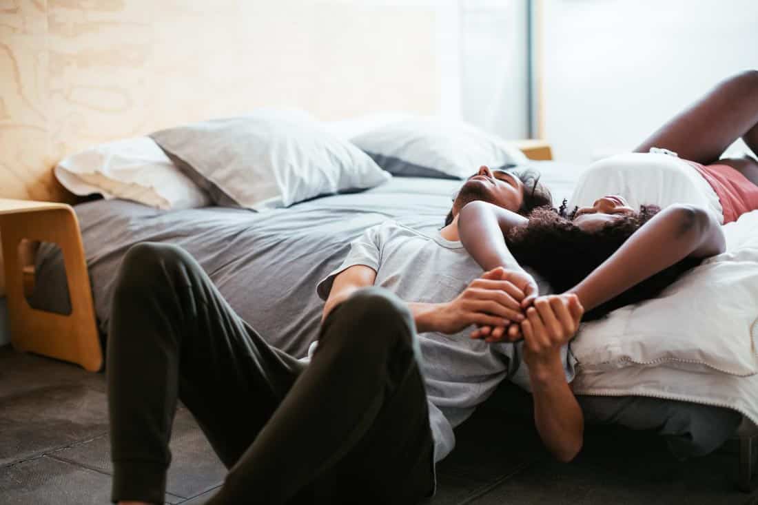 Man sitting on floor while lying back on bed holding a woman's hands, representing affair counseling