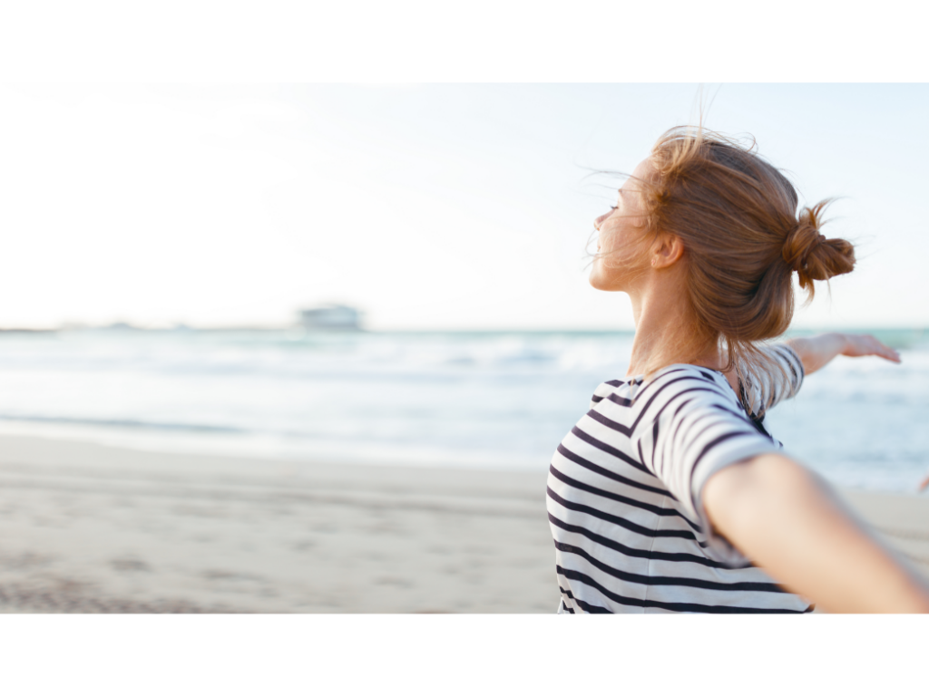 A woman stands by the ocean spreading her arms wide representing how to get unstuck when you're feeling trapped.