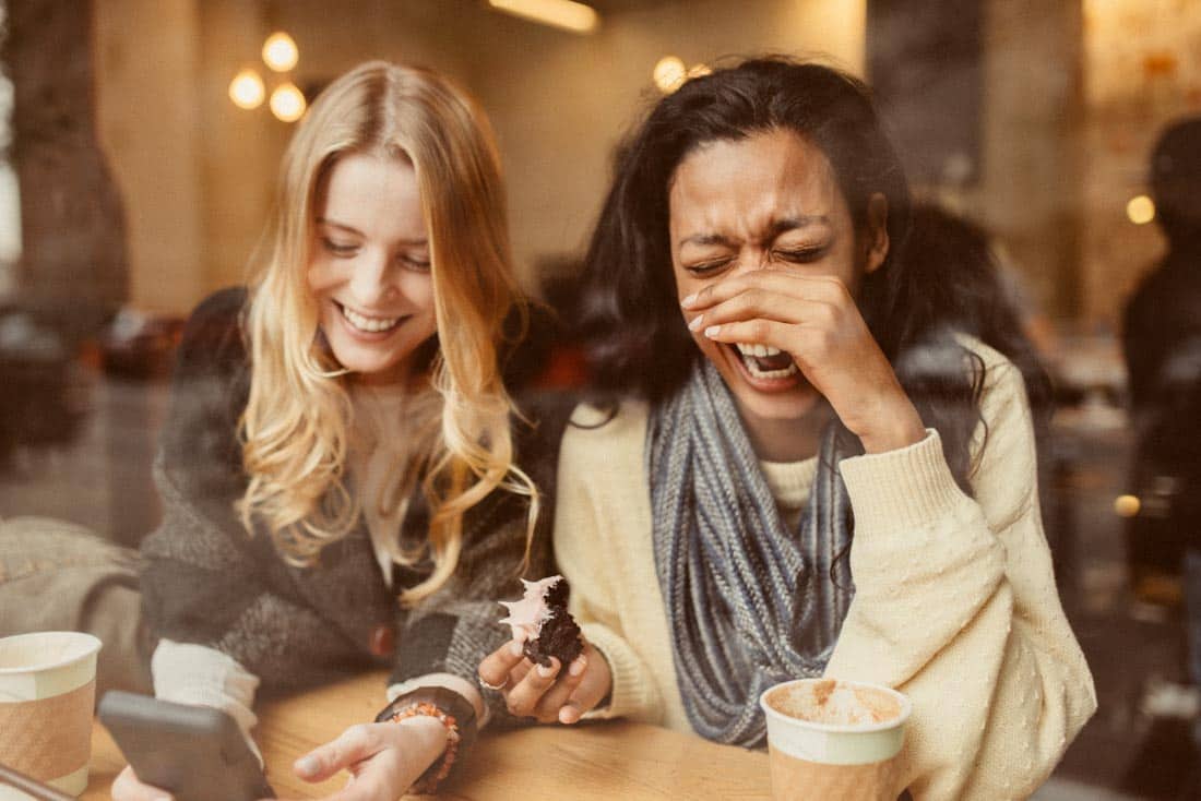 Two women laughing in coffee shop showing authentic happiness and emotional intelligence