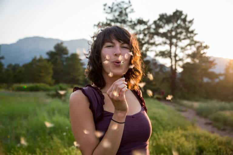 Denver Life Coaching: Woman in the mountain blowing a dandelion into the camera