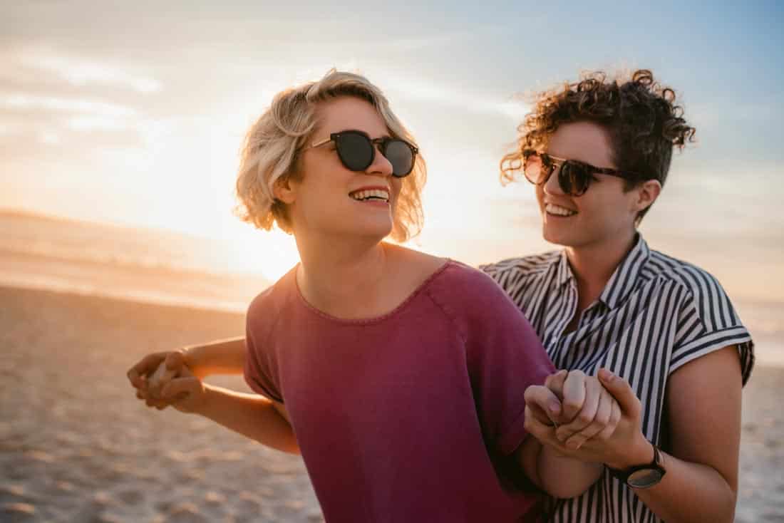 Lesbian couple holding hands on beach at sunset.