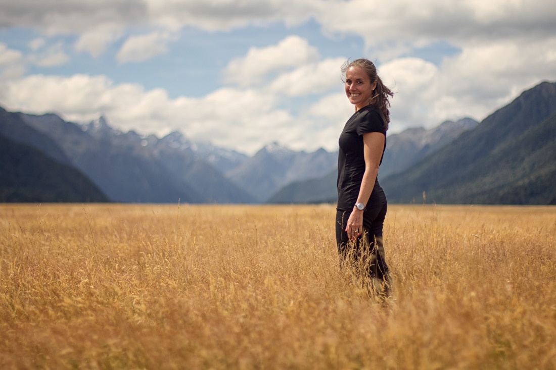 Achieve personal growth: a young pretty girl stands in a field of tall gold grass / wheat with a backdrop of mountains and a cloudy blue sky. 