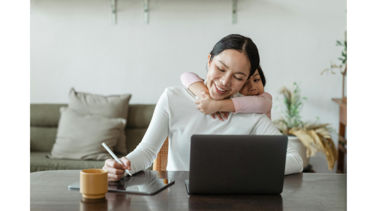A smiling woman works at a laptop while a child hugs her representing emotional intelligence as the key to love, happiness and success