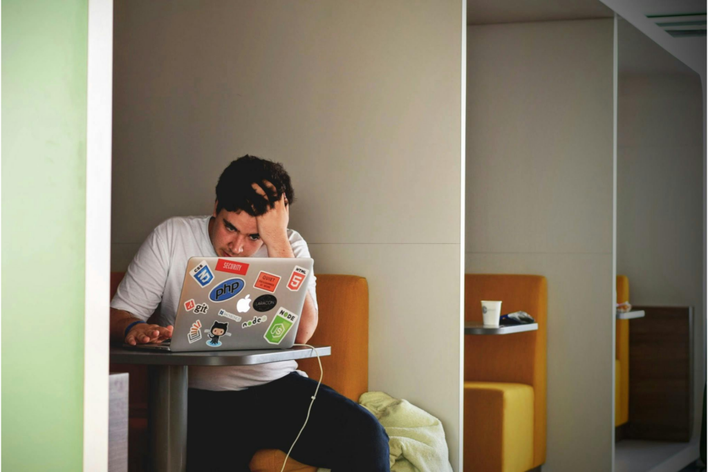 A man working on a laptop holds his head in his hands representing the signs of a toxic boss