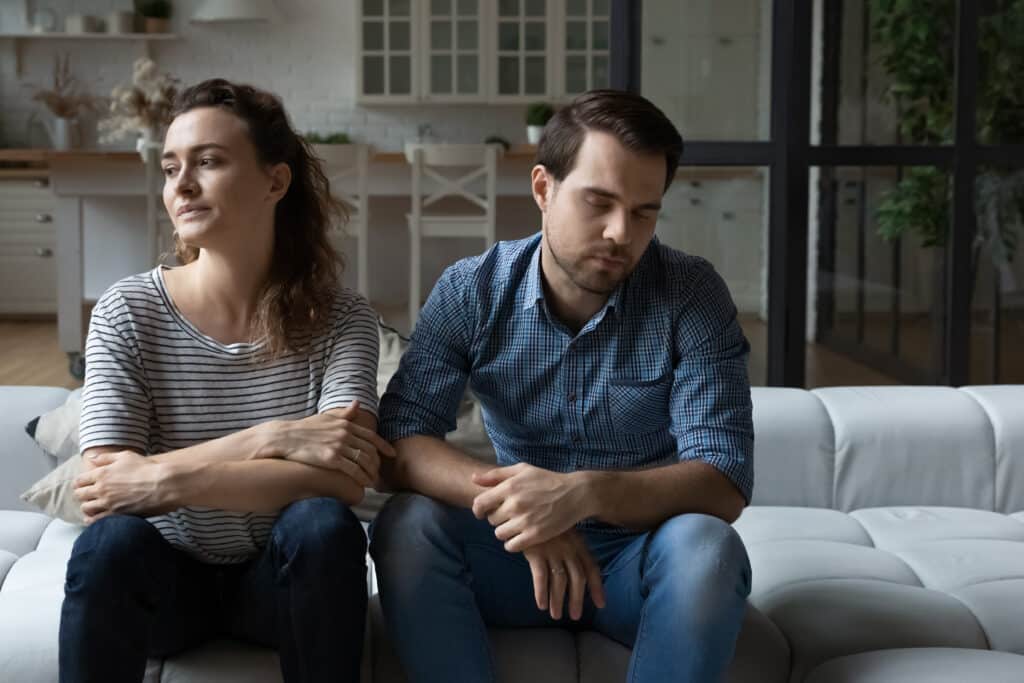 A woman and man sit on their couch looking tired and unhappy representing Why the “relationships are hard work” idea is just wrong.