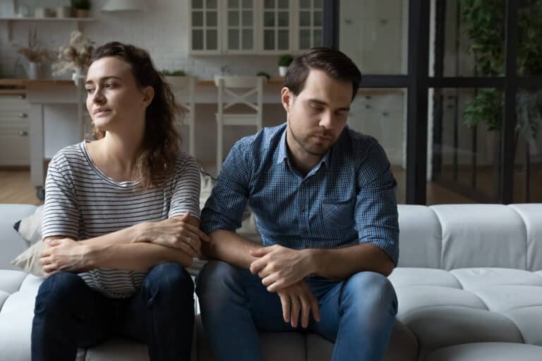 A woman and man sit on their couch looking tired and unhappy representing Why the “relationships are hard work” idea is just wrong.