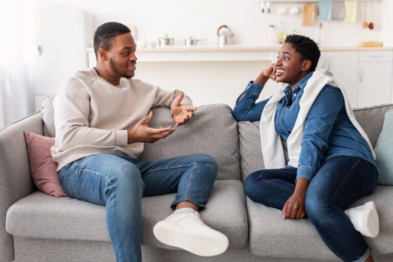 A black man and woman sit on the couch having a conversation representing How to Be a Better Communicator.