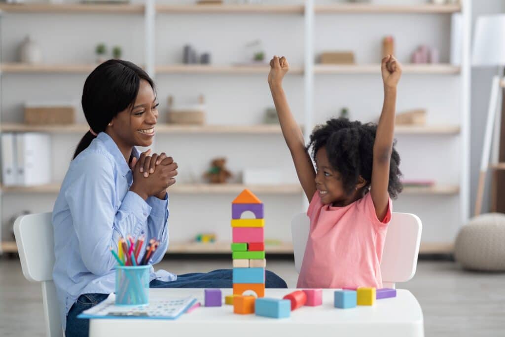 A woman watches her child build a tower with blocks and the child stretches her arms up in celebration as she finishes representing child centered play therapy.