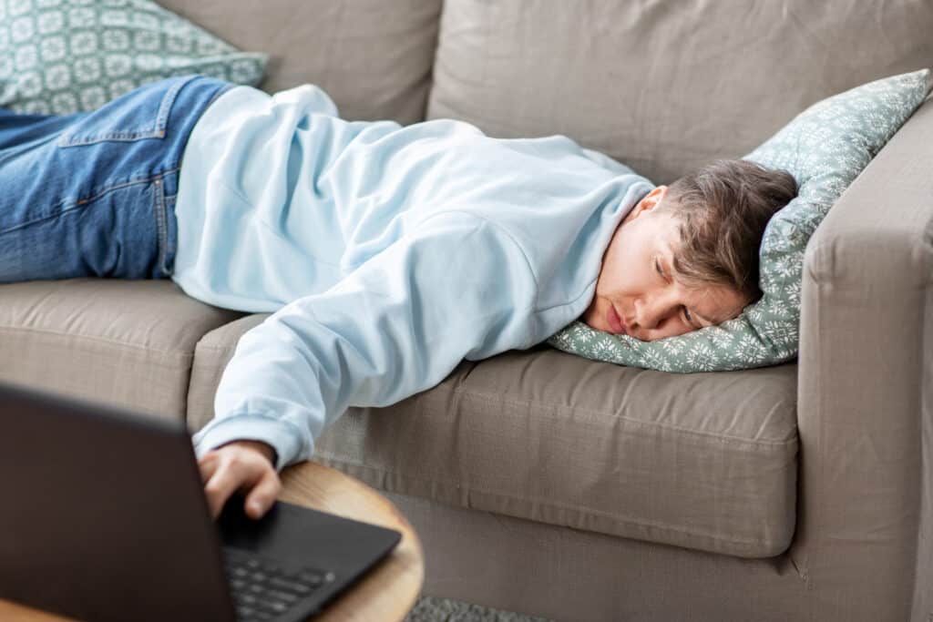 A man lies slumped on the couch watching tv on his laptop representing how to get out of a slump.