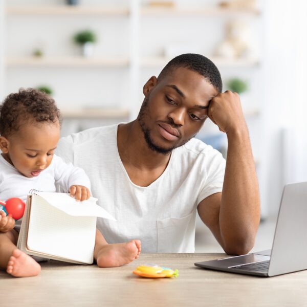 A man sits at a table in his home working from his laptop and taking care of his infant at the same time. He looks burned out representing Parental Burnout: How to Find Work-Life Balance as a Parent