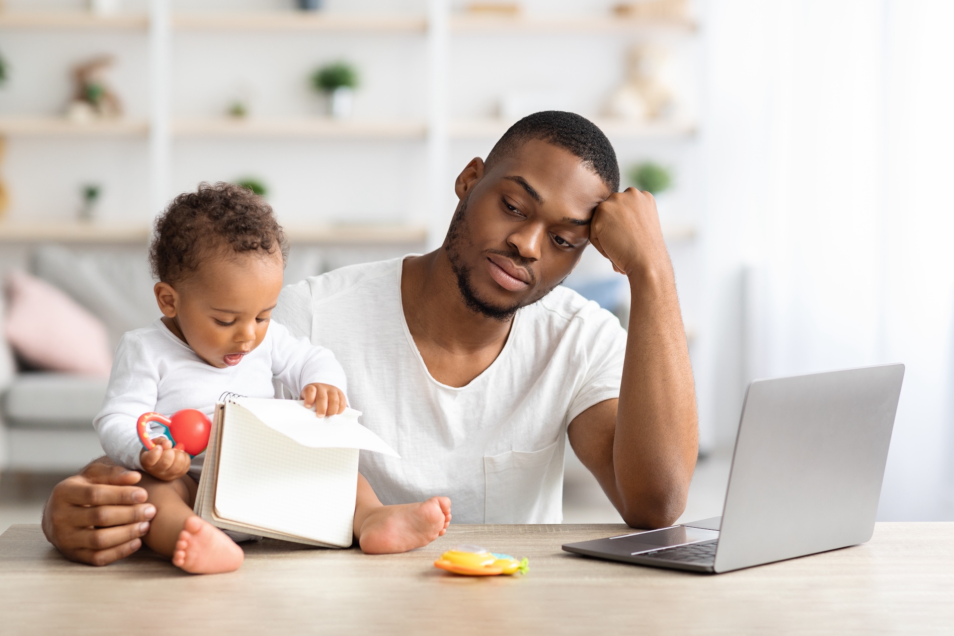 A man sits at a table in his home working from his laptop and taking care of his infant at the same time. He looks burned out representing Parental Burnout: How to Find Work-Life Balance as a Parent
