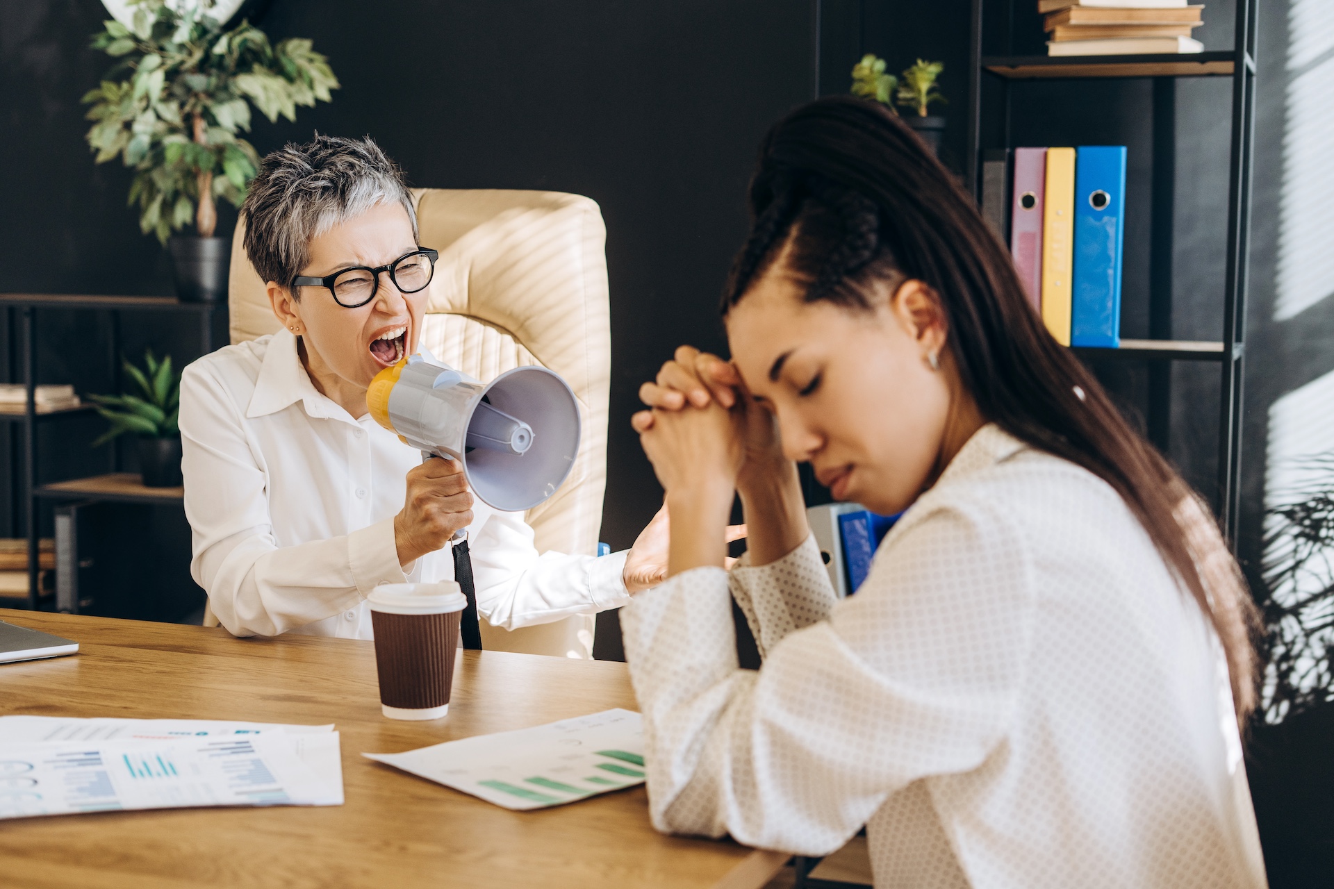 Employee stressed at desk while bully boss yells through megaphone in toxic workplace