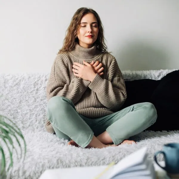 Woman sitting cross-legged on a couch with eyes closed and hands on chest, practicing mindful self-compassion and emotional healing.