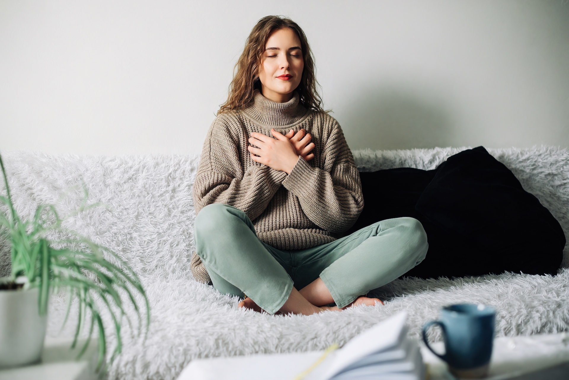 Woman sitting cross-legged on a couch with eyes closed and hands on chest, practicing mindful self-compassion and emotional healing.