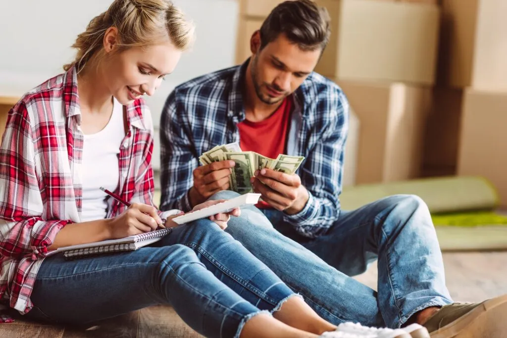 A couple sitting on the floor planning their finances together, counting money and taking notes — symbolizing money and relationships and combining finances after marriage.