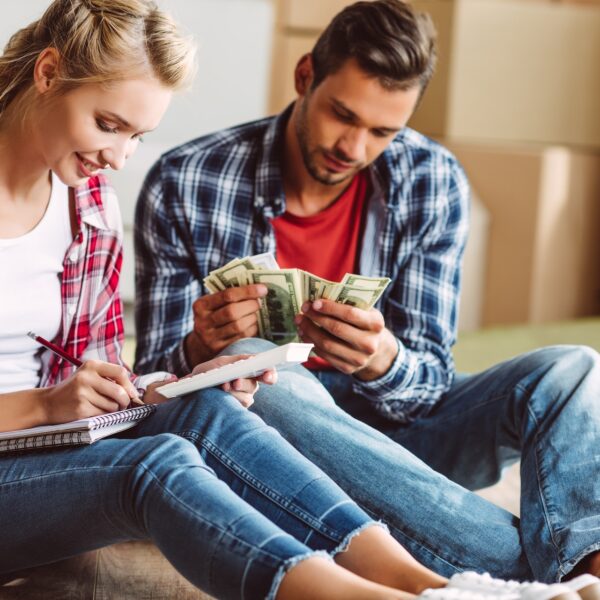 A couple sitting on the floor planning their finances together, counting money and taking notes — symbolizing money and relationships and combining finances after marriage.
