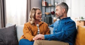 A couple sitting on a couch holding hands and having a serious conversation, representing communication breakdown and relationship repair.