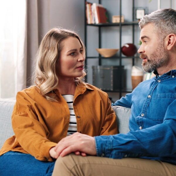 A couple sitting on a couch holding hands and having a serious conversation, representing communication breakdown and relationship repair.