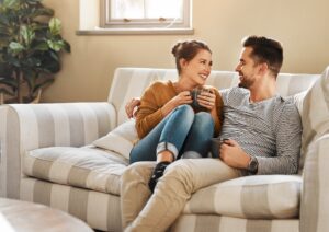 A couple sitting close together on a couch, smiling and talking while holding coffee, illustrating attentive listening and emotional connection in a relationship.