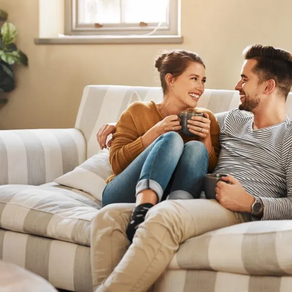 A couple sitting close together on a couch, smiling and talking while holding coffee, illustrating attentive listening and emotional connection in a relationship.