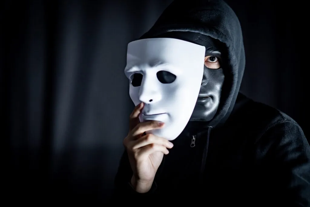 A masked figure holding a white face mask, symbolizing a con artist hiding their true identity through deception and emotional manipulation.