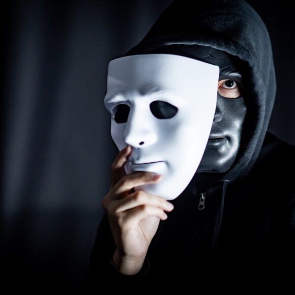 A masked figure holding a white face mask, symbolizing a con artist hiding their true identity through deception and emotional manipulation.