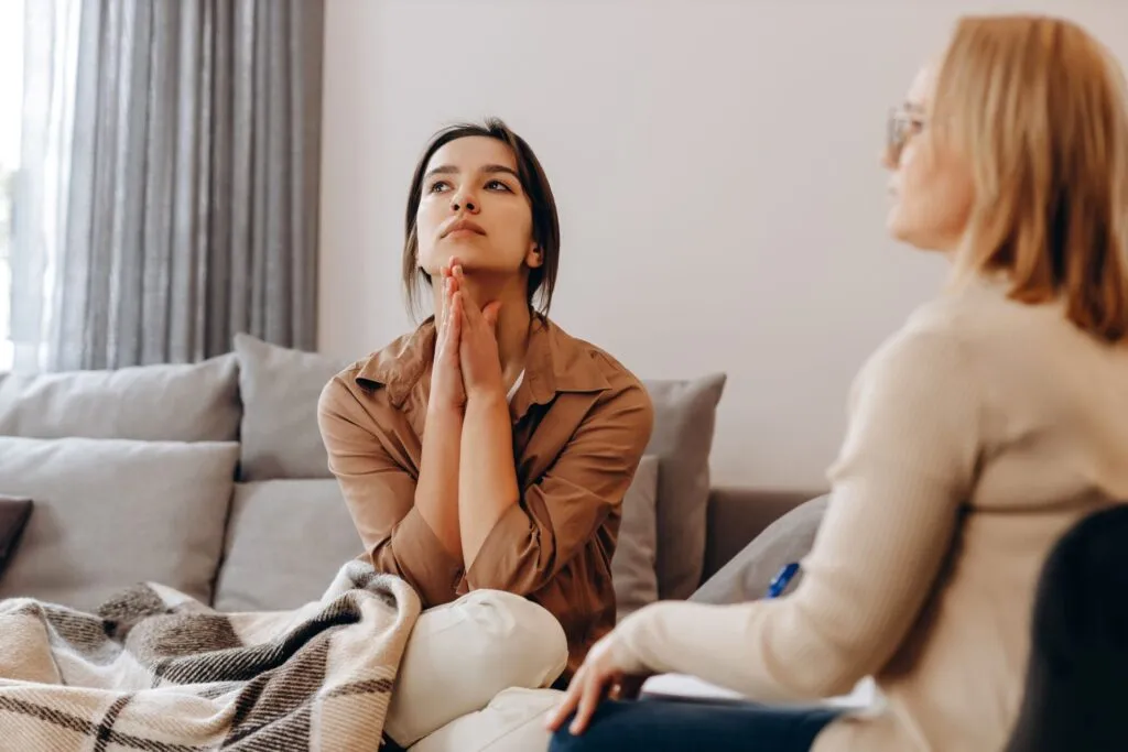 Client sits on a couch with hands clasped under her chin, looking distressed and disappointed during a therapy session.