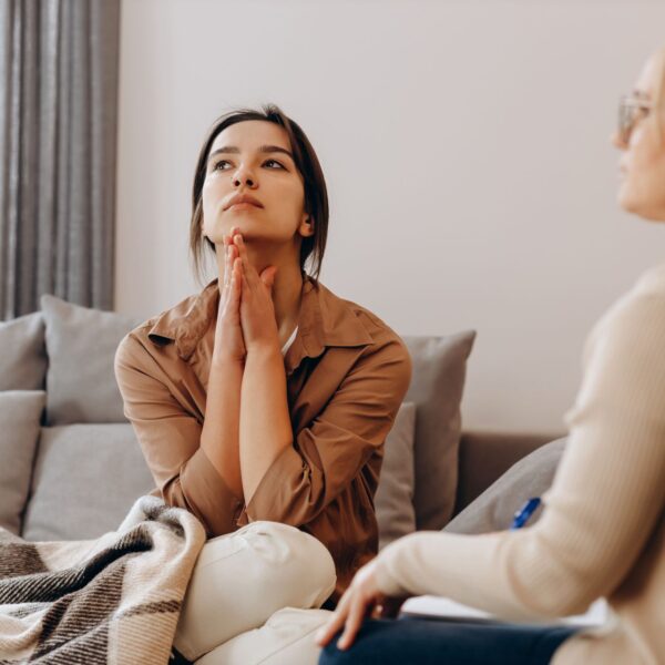 Client sits on a couch with hands clasped under her chin, looking distressed and disappointed during a therapy session.