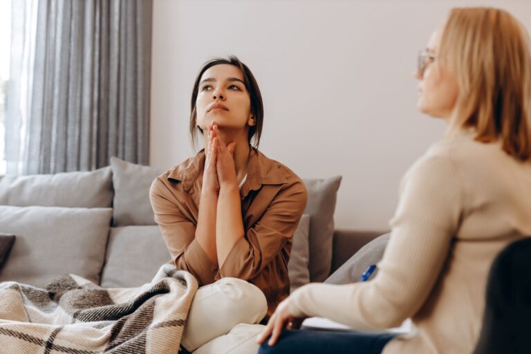 Client sits on a couch with hands clasped under her chin, looking distressed and disappointed during a therapy session.