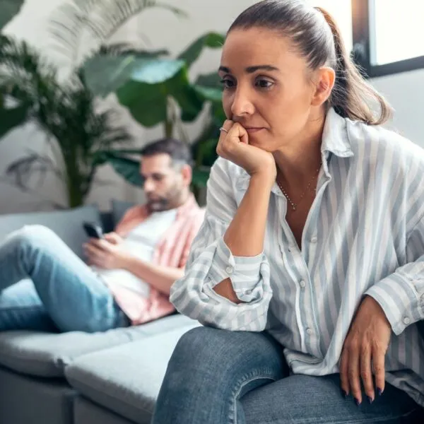 A woman looking worried while partner sits on the couch using phone in the background, illustrating feeling lonely in marriage and an emotionally checked-out spouse.