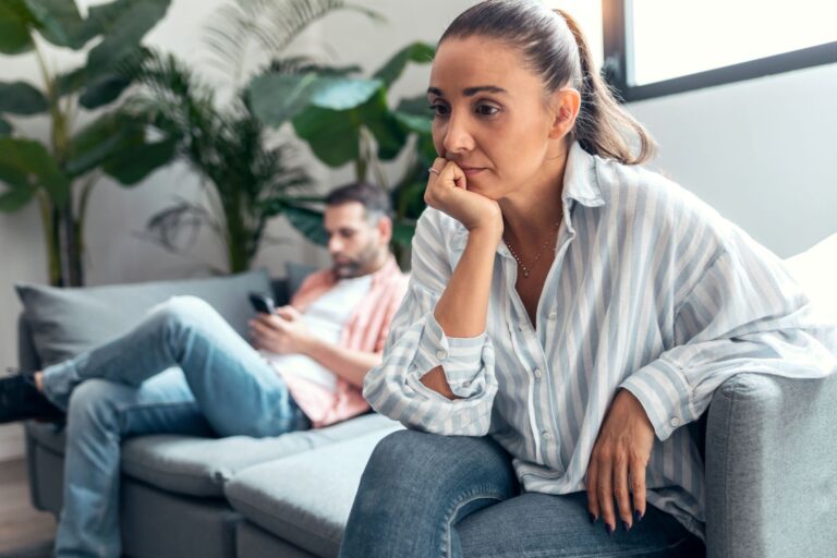 A woman looking worried while partner sits on the couch using phone in the background, illustrating feeling lonely in marriage and an emotionally checked-out spouse.