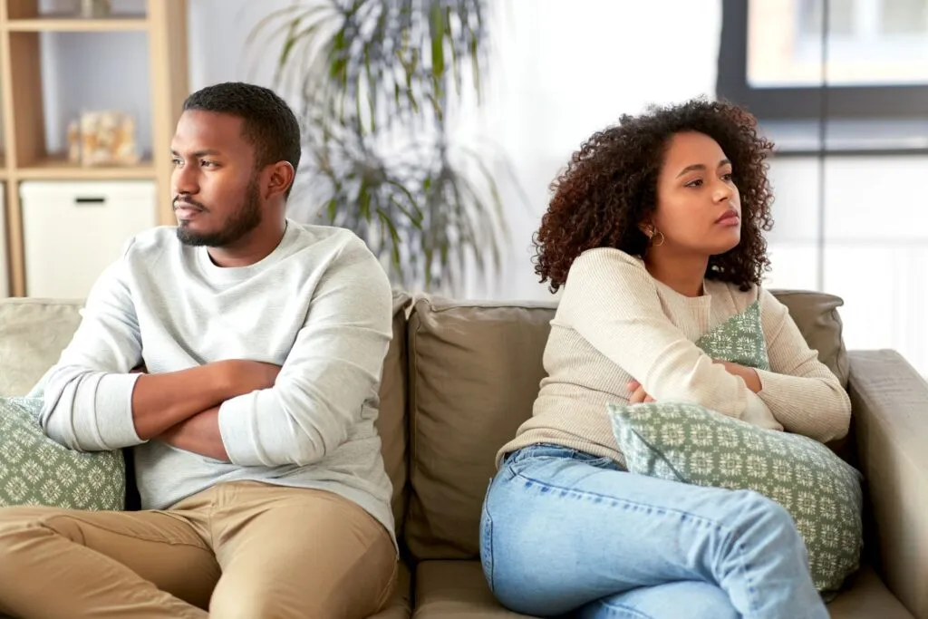Couple sitting back-to-back on a couch with arms crossed, illustrating the power struggle stage in relationships and emotional disconnection during conflict.