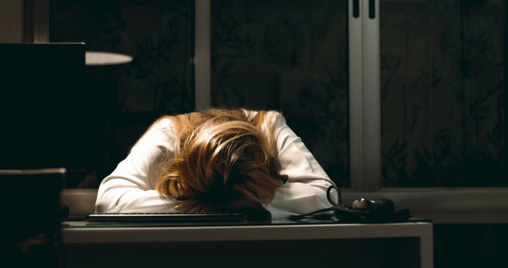 Woman with head down on desk at night, exhausted from stress and burnout while working on a computer.