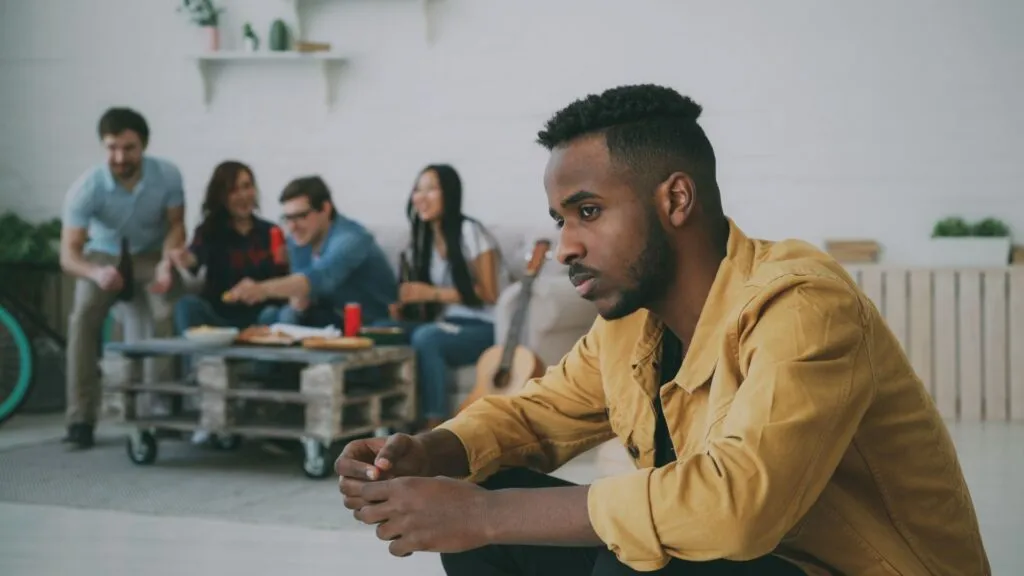 Man sitting apart from a group of friends looking thoughtful, illustrating loneliness and the challenge of making friends as an adult.