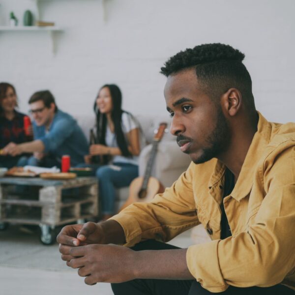 Man sitting apart from a group of friends looking thoughtful, illustrating loneliness and the challenge of making friends as an adult.