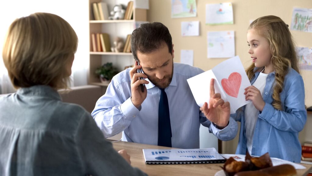 Father on a work phone call at home while his daughter shows him a drawing, illustrating the need to set boundaries at work and protect personal life