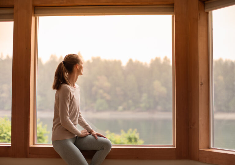 Woman sitting by a large window, looking out at a hazy landscape, reflecting climate anxiety and uncertainty.