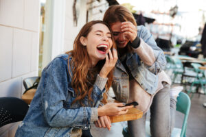Two women laughing together at an outdoor café, sharing a playful moment over a phone.