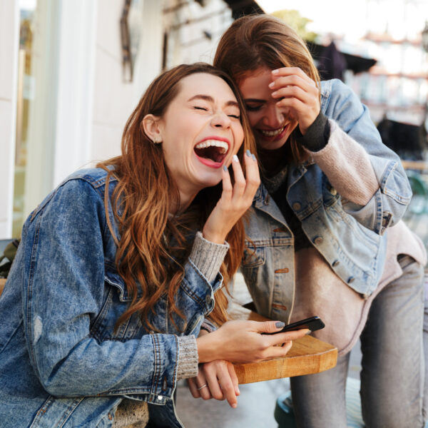 Two women laughing together at an outdoor café, sharing a playful moment over a phone.