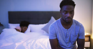 Man sitting on the edge of a bed looking distressed while his partner lies blurred in the background, suggesting relationship conflict and emotional distance.