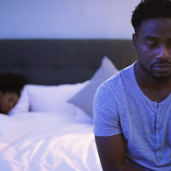 Man sitting on the edge of a bed looking distressed while his partner lies blurred in the background, suggesting relationship conflict and emotional distance.