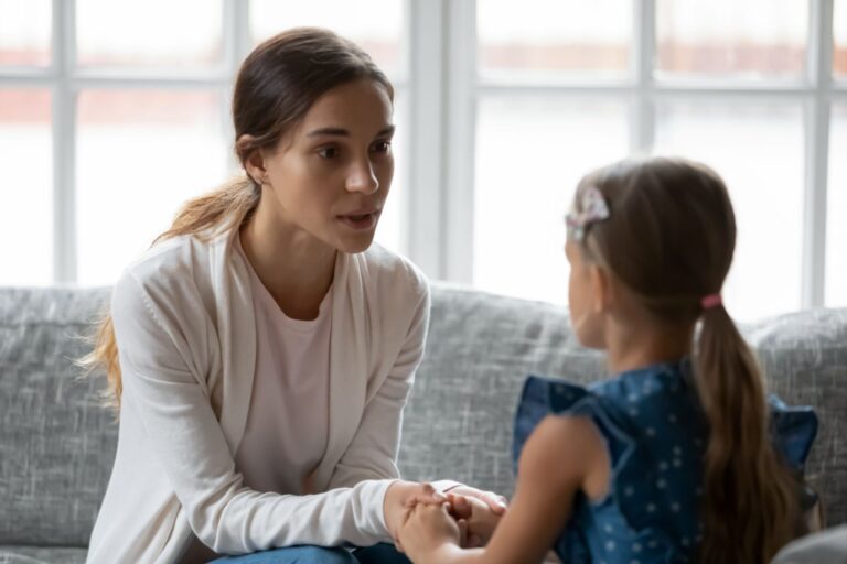 Parent holding a child’s hands during a calm conversation on a couch, illustrating emotion coaching and secure attachment parenting.